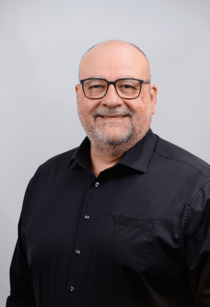 Porträtfoto eines Mannes mit Bart und Brille, im dunklen Hemd.